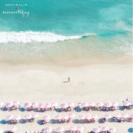 Aerial beach scene with turquoise ocean waves, pale sand, rows of pink-and-white umbrellas, and a small person walking near the shoreline. The text 'GRAYMALIN BESPOKE Wedding' appears in the upper left.
