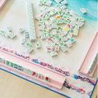 A pink Mahjong game set with white tiles featuring multicolor Chinese characters and symbols, arranged on a decorative mat on a light wood surface.