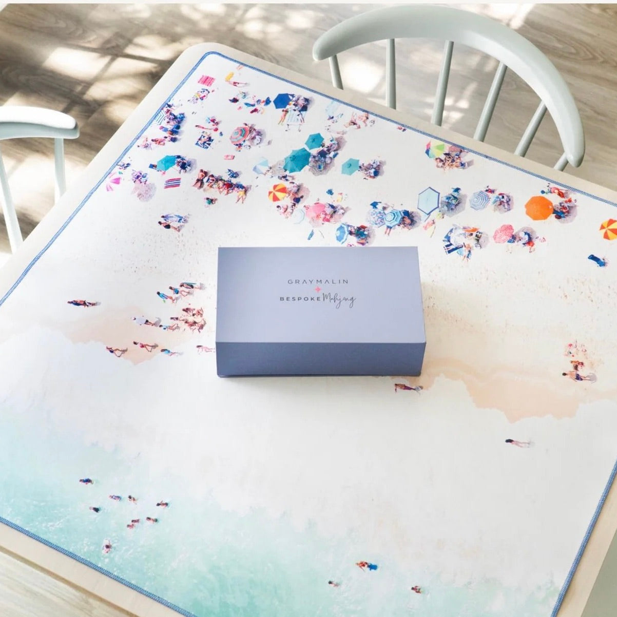 A square gray box with the text 'GRAYMALIN x MEMENTO MEMENTO' sits on a large beach-themed table mat. The mat features a watercolor-style design with a light blue and beige shoreline and small colorful beachgoers.