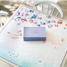 A square gray box with the text 'GRAYMALIN x MEMENTO MEMENTO' sits on a large beach-themed table mat. The mat features a watercolor-style design with a light blue and beige shoreline and small colorful beachgoers.