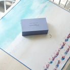 A square gray box with the text 'GRAYMALIN x RESPONSE Design' sits on a large printed mat or sheet featuring a light blue watercolor wash on the left and a row of pink beach umbrellas on the right, photographed from above on a light wood surface.