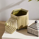 Two decorative ceramic containers with striped patterns, one hexagonal open jar and one square lidded box, displayed on a white surface against a light neutral background.