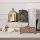 Decorative home accessories arranged on a shelf, including a striped ceramic lidded box, a horse-shaped sculpture, books, small animal figurines, and a rectangular striped tray, against a white paneled wall.