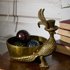 A decorative brass bird figurine in the shape of a rooster wearing a small top hat, placed beside a matching brass bowl filled with colorful ornaments on a wooden surface.
