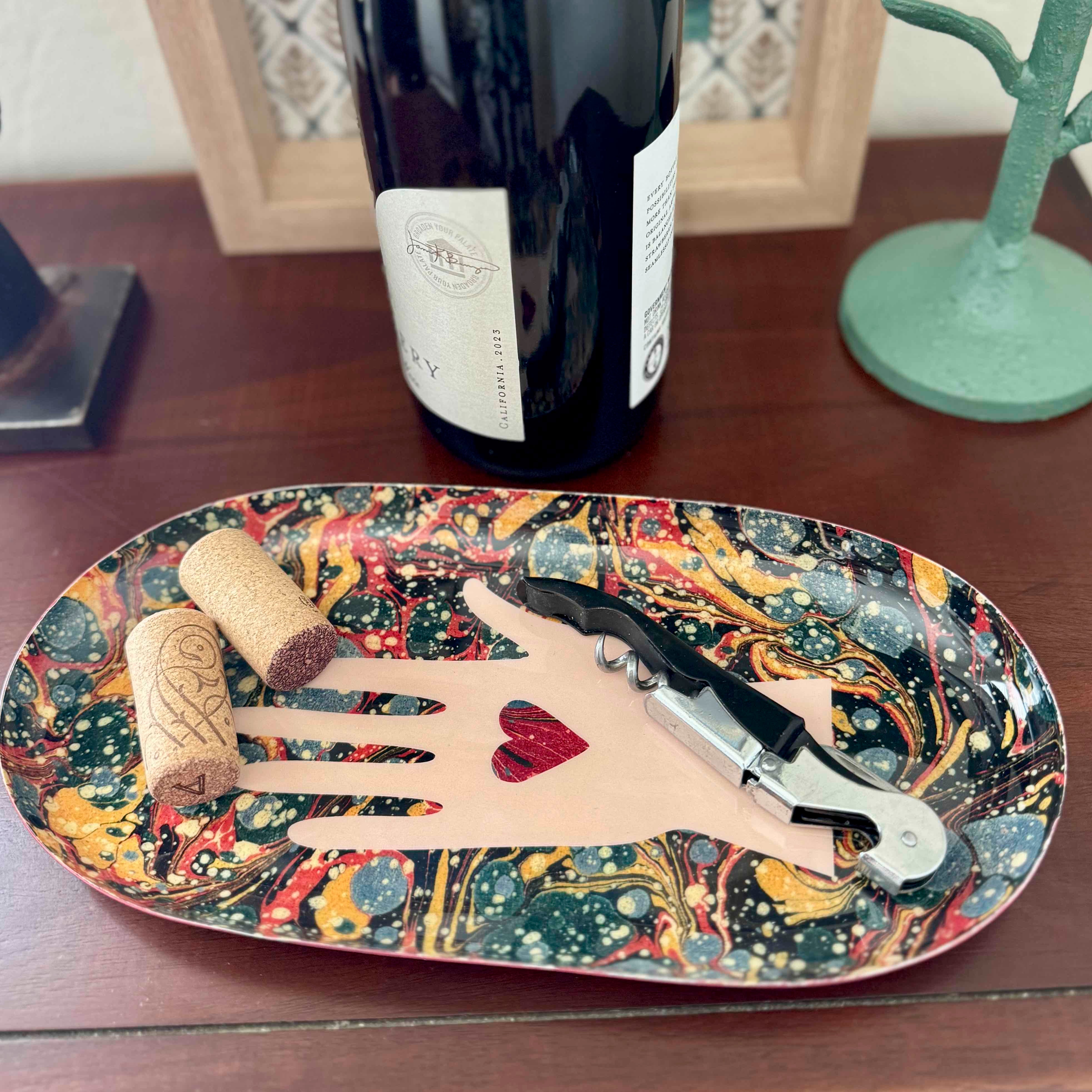 Decorative tray with a hand design holding a corkscrew and cork, next to a bottle of wine.