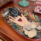 Decorative tray with a hand holding a heart, key fob, and coins on a wooden surface.