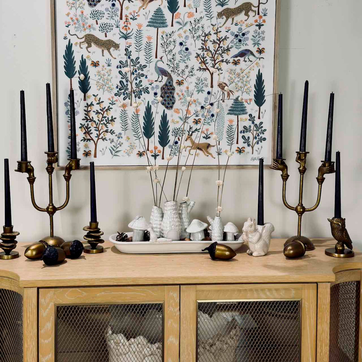 A styled interior scene showing a light wood sideboard with black taper candles in brass and bronze candelabras, white ceramic vases with dried stems, mushrooms, acorns, and small decorative figurines, and a large framed forest illustration hanging on the wall behind.