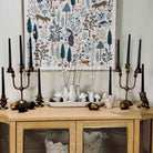 A styled interior scene showing a light wood sideboard with black taper candles in brass and bronze candelabras, white ceramic vases with dried stems, mushrooms, acorns, and small decorative figurines, and a large framed forest illustration hanging on the wall behind.