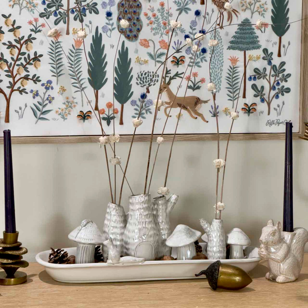 A styled tabletop arrangement featuring a white ceramic tray with textured white mushroom-shaped vases holding dried stems, flanked by dark blue taper candles in gold and white holders. A white ceramic squirrel figurine and a gold acorn sit in the foreground, set against a light wood wall and a large forest-themed wall hanging.