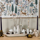 A styled tabletop arrangement featuring a white ceramic tray with textured white mushroom-shaped vases holding dried stems, flanked by dark blue taper candles in gold and white holders. A white ceramic squirrel figurine and a gold acorn sit in the foreground, set against a light wood wall and a large forest-themed wall hanging.