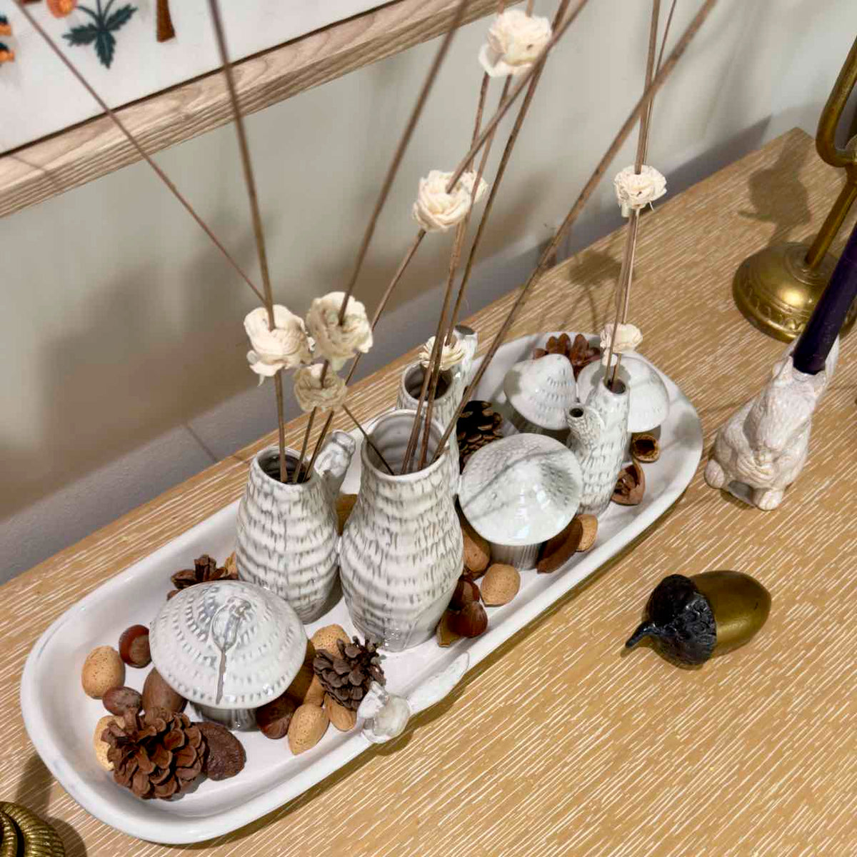 A decorative white ceramic tray holding several textured white vases with dried flower stems, arranged on a light wood surface with pinecones and small decorative objects around it.