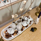 A decorative white ceramic tray holding several textured white vases with dried flower stems, arranged on a light wood surface with pinecones and small decorative objects around it.
