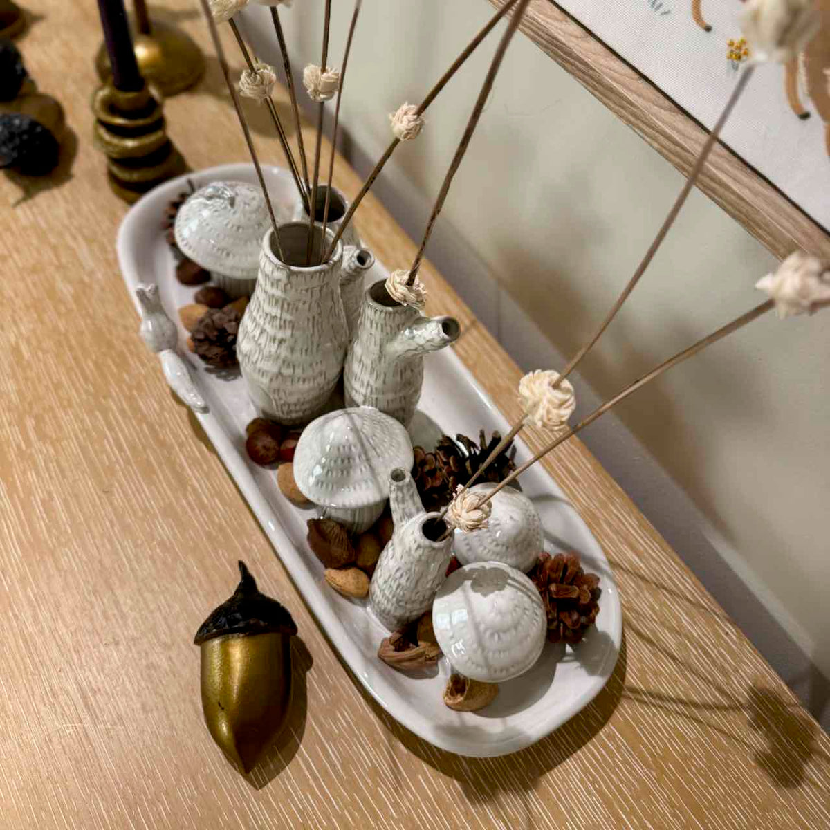 A decorative ceramic bird and mushroom tray arrangement on a wooden table, styled with dried stems, pinecones, acorns, and a gold acorn ornament.
