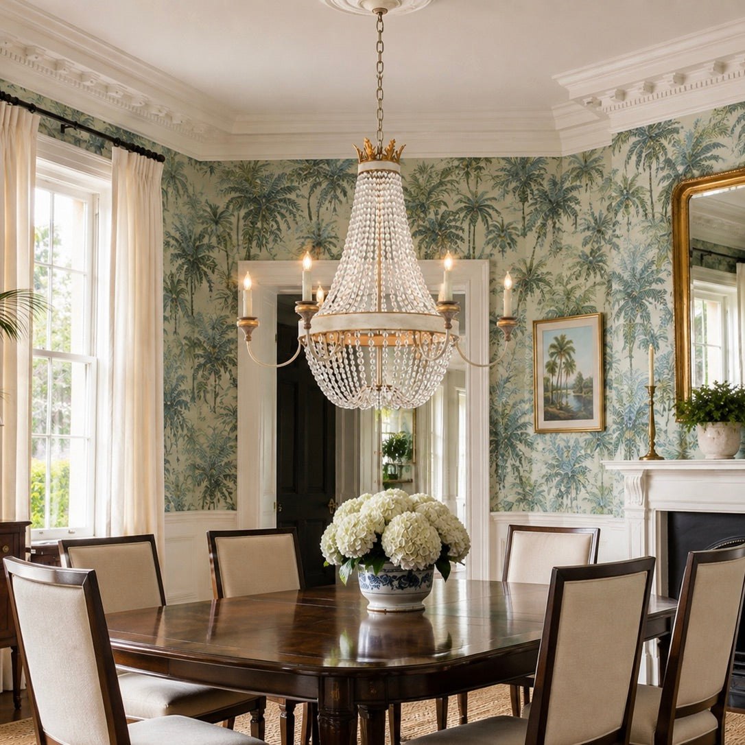An elegant dining room featuring a large crystal chandelier hanging over a dark wood dining table with upholstered chairs. The room is decorated with green and white palm tree pattern wallpaper, white walls with molding, and a fireplace with a gold mirror.