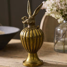 A decorative metallic rabbit-shaped container with a ribbed bulbous body and pedestal base, shown on a wooden table with blurred flowers and home decor in the background.