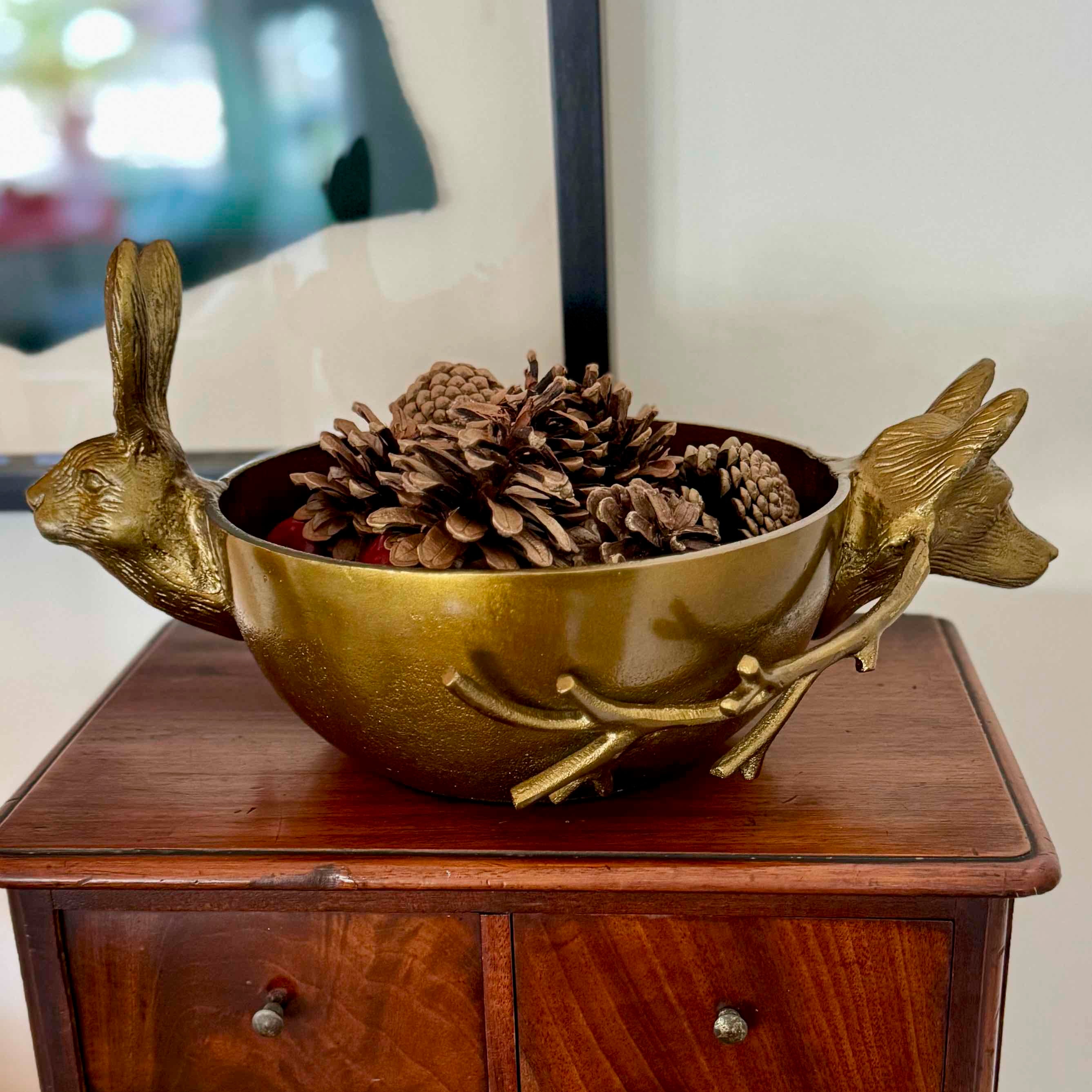 Decorative gold bowl with rabbit handles filled with pinecones on a wooden surface.