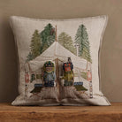 A square beige linen throw pillow featuring an embroidered winter camping scene with a tent, pine trees, and two figures in colorful parkas, displayed on a wooden surface against a neutral wall.