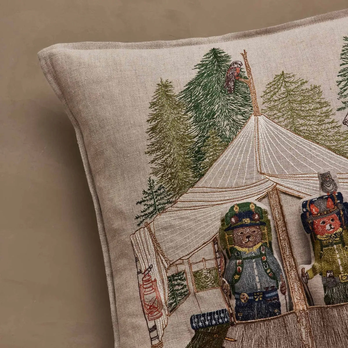 Close-up of a beige linen throw pillow featuring embroidered camping scene with a canvas tent, pine trees, and two bear characters in winter clothing.