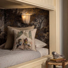 A styled bedroom corner featuring a bed with a decorative throw pillow printed with a vintage camping scene. The background is decorated with dark, moody botanical wallpaper and a warm wall sconce.