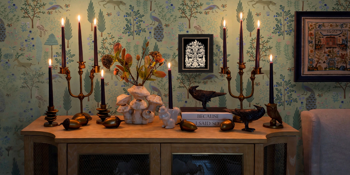 A styled interior scene showing a wooden console table against patterned wallpaper, decorated with multiple black and bronze candelabras, lit candles, sculptural bird figures, a white shell arrangement, and framed wall art.