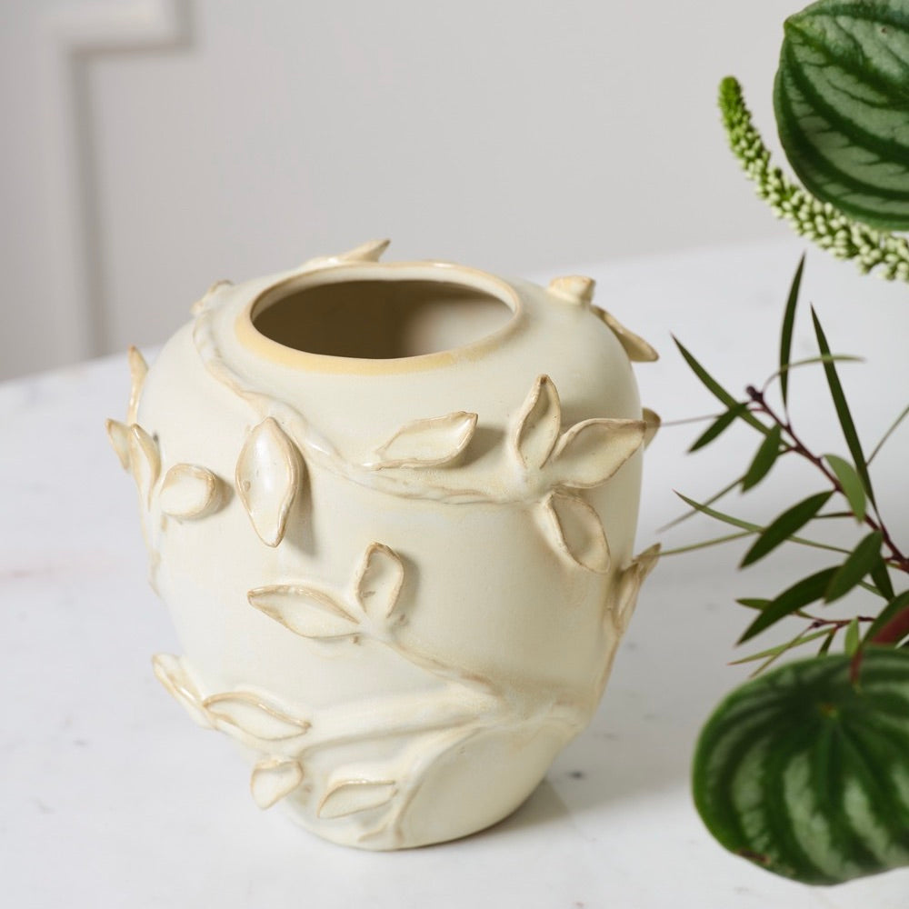 An off-white ceramic bud vase with raised leaf and branch details, styled on a white marble surface next to green houseplants.