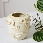 An off-white ceramic bud vase with raised leaf and branch details, styled on a white marble surface next to green houseplants.