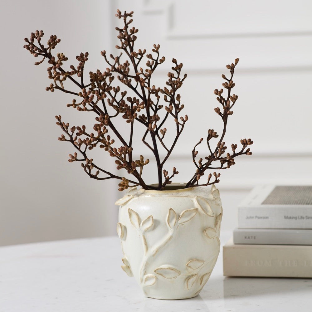 A cream-colored ceramic vase with embossed leaf patterns, holding dried brown branches, styled on a white marble surface next to stacked books.