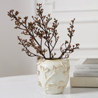 A cream-colored ceramic vase with embossed leaf patterns, holding dried brown branches, styled on a white marble surface next to stacked books.