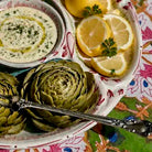 A close-up of a serving platter holding two green artichokes, a small bowl of white dip with herbs and olive oil, and lemon slices on a patterned tablecloth.