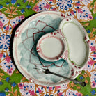 A ceramic serving platter with three sections, featuring a central bowl, a small oval dish, and a larger oval compartment, decorated with a green and pink floral pattern. A small metal serving fork rests on the platter, which is set against a colorful floral patterned fabric background.