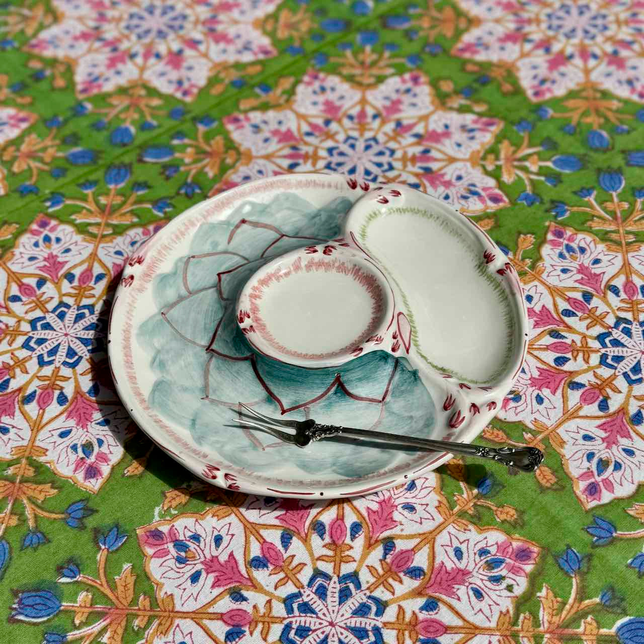 A ceramic three-compartment serving plate with a matching spoon, displayed on a vibrant, patterned green tablecloth.