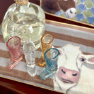 Clear bottle with a gold cap on a tray with colorful shot glasses and a cow painting in the background.