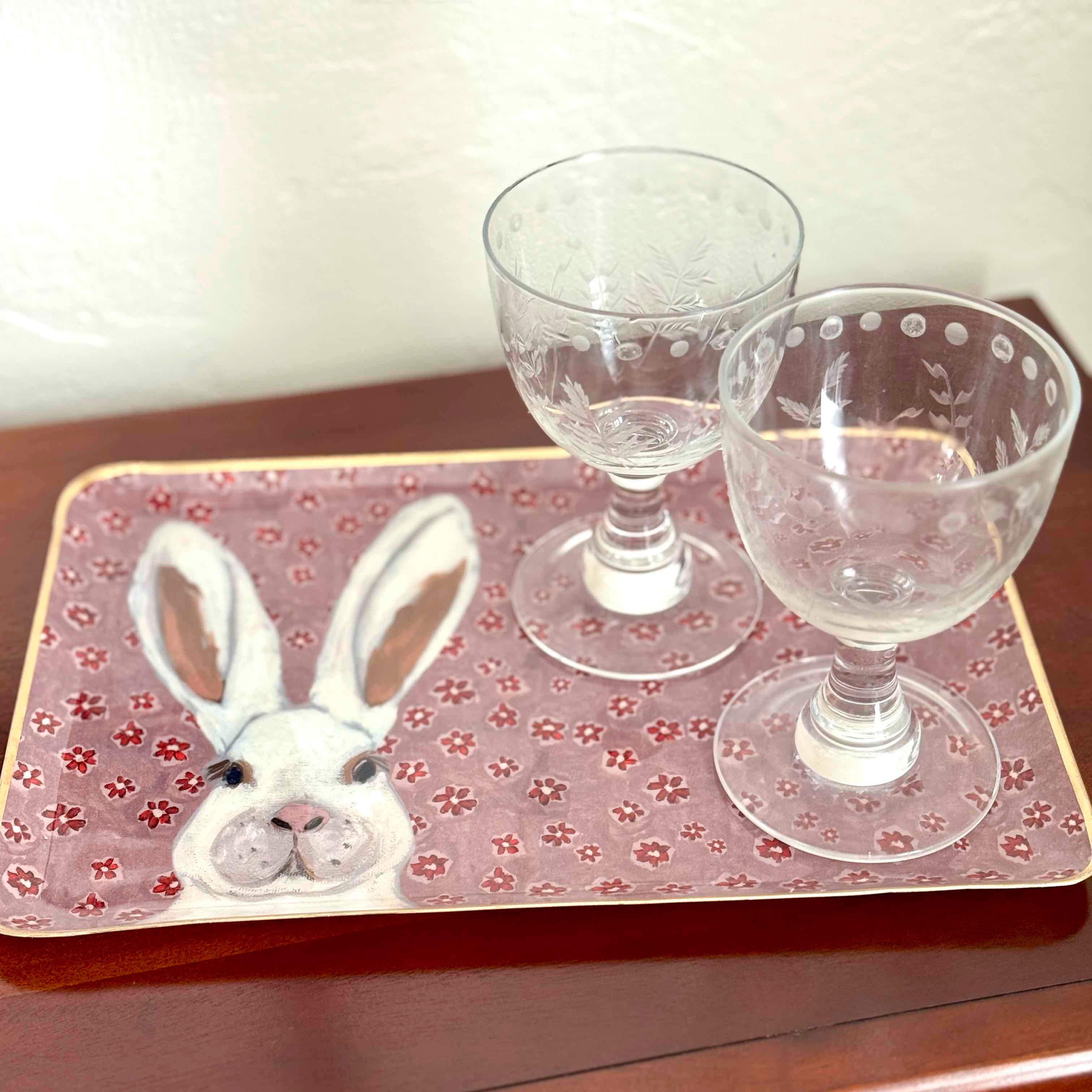 Two glasses on a decorative tray with a rabbit design on a wooden surface.