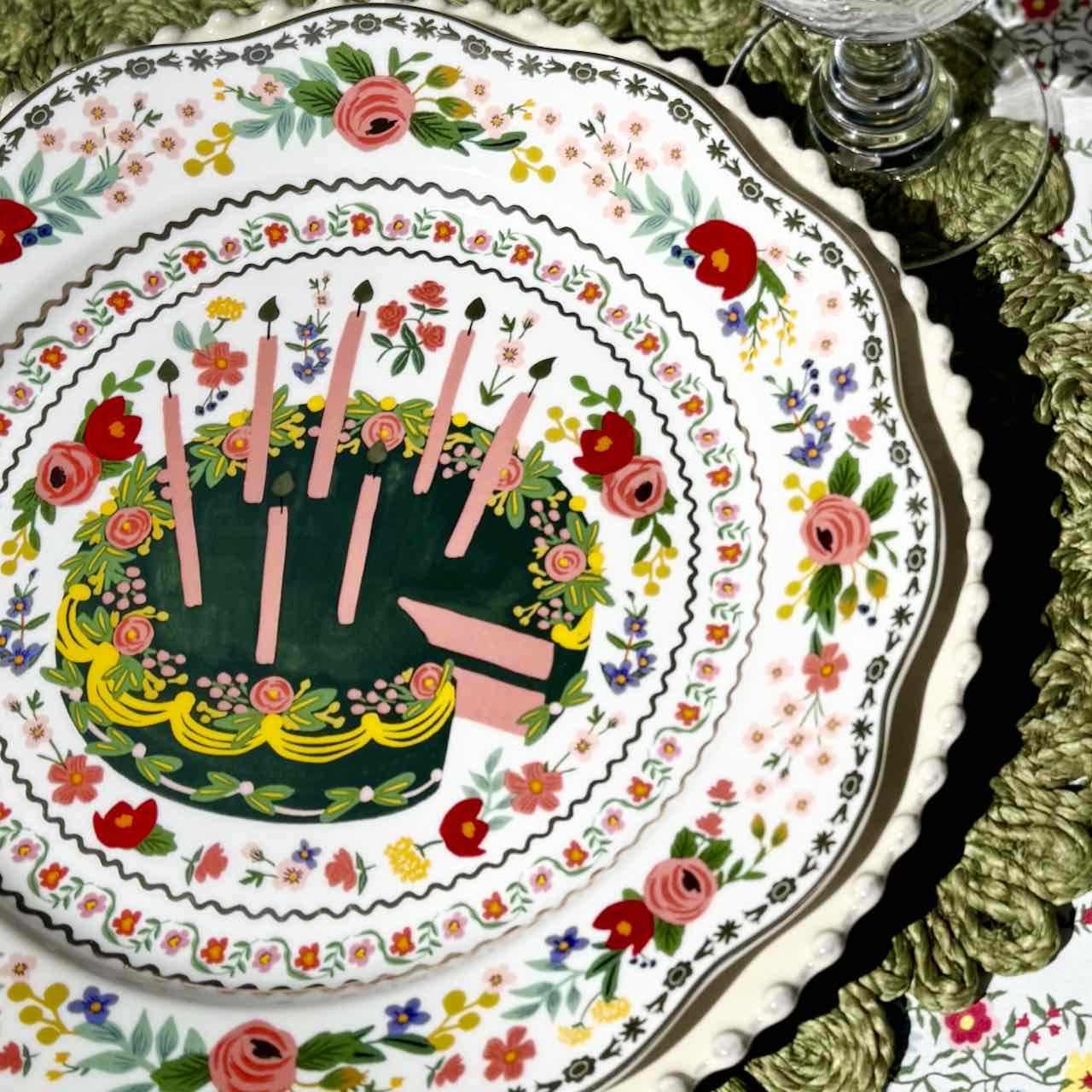 Decorative plate with a cake design featuring candles and flowers on a floral-patterned tablecloth.
