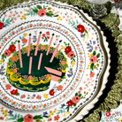 Decorative plate with a cake design featuring candles and flowers on a floral-patterned tablecloth.