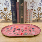 Pink decorative tray with floral designs on a wooden surface with books in the background