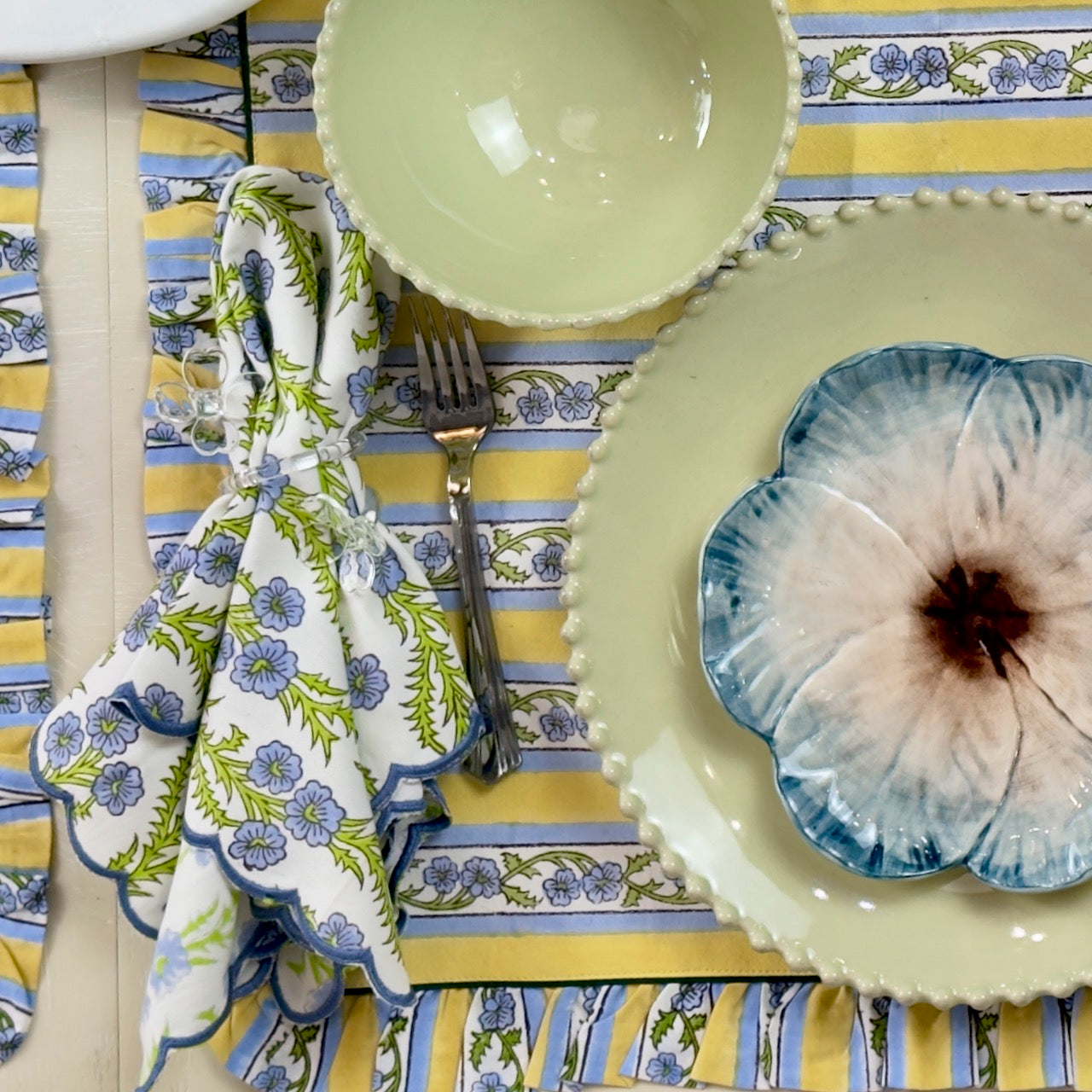 Table setting with floral tablecloth, plates, and cutlery on a yellow and blue striped tablecloth.