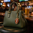 An olive green textured leather handbag with gold-tone hardware sits on a black leather stool in a bar setting. The bag features a football-shaped keychain charm and is positioned in front of a blurred bar background with a glass of beer.