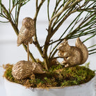Three metallic gold garden figurines of an owl, a hedgehog, and a squirrel arranged on moss and branches in a white planter.