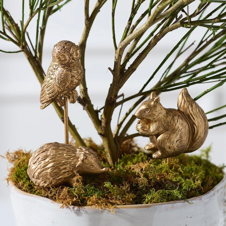 Three metallic gold garden figurines of an owl, a hedgehog, and a squirrel arranged on moss and branches in a white planter.