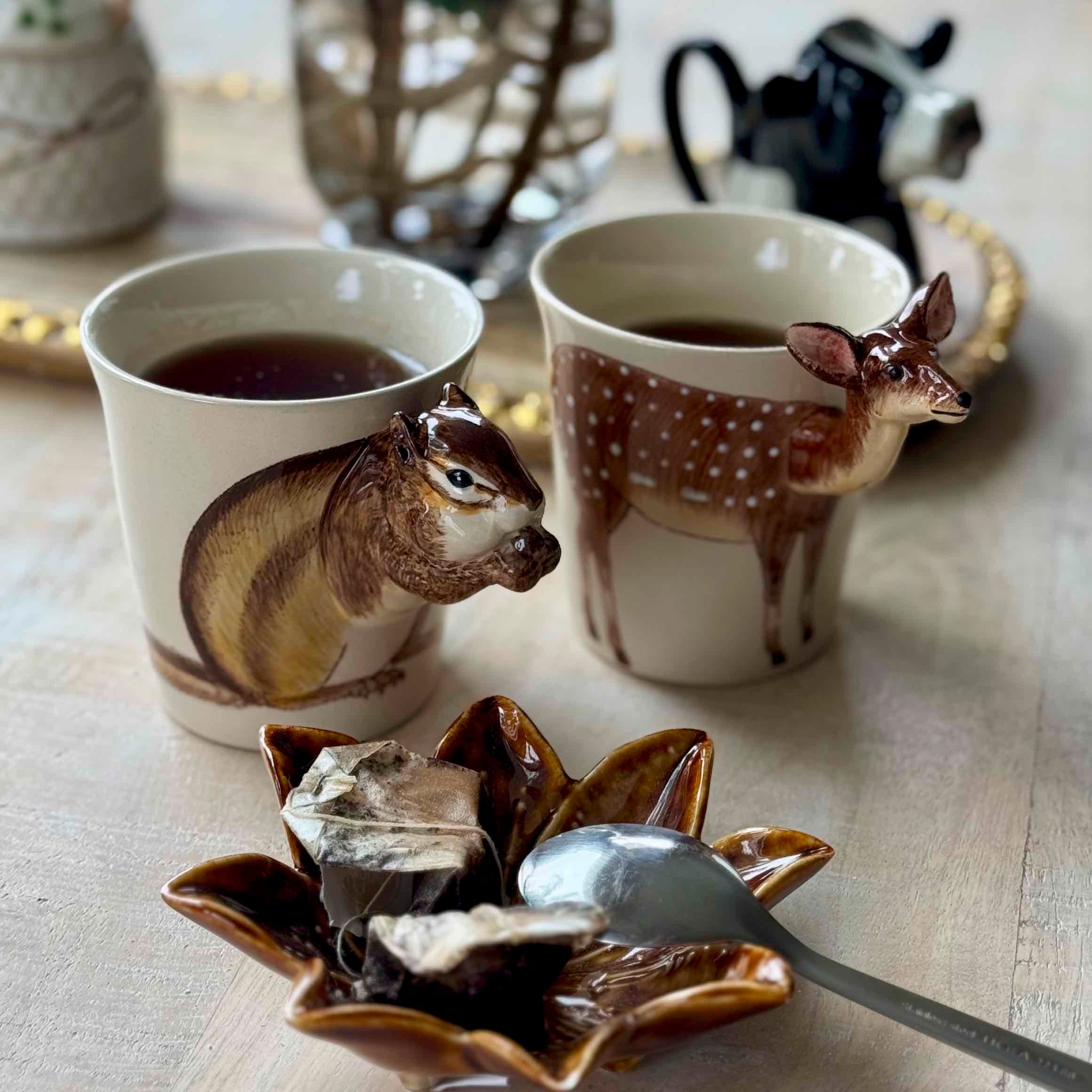 Two ceramic cups with animal designs, a spoon, and tea bags on a wooden surface.