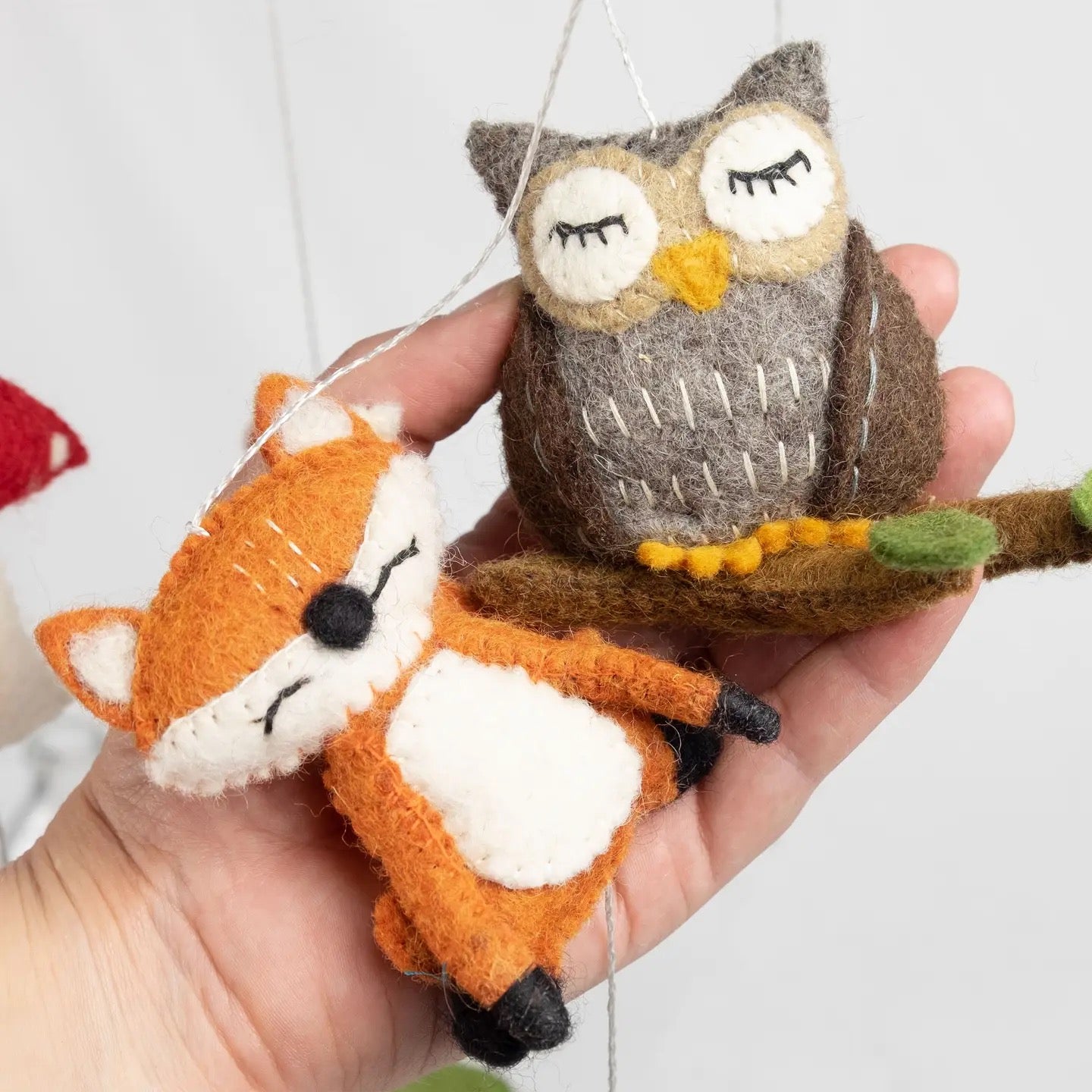 Hand holding two felt animal ornaments, an owl and a fox, on a white background.