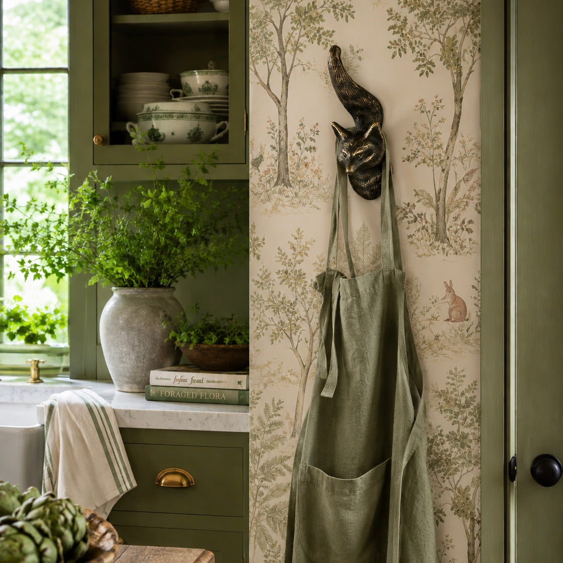 A sage green linen apron hangs from a dark metal cat-shaped hook on a wall with beige botanical wallpaper. The scene is a styled kitchen corner with olive green cabinets, a window, and potted greenery.