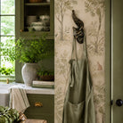 A sage green linen apron hangs from a dark metal cat-shaped hook on a wall with beige botanical wallpaper. The scene is a styled kitchen corner with olive green cabinets, a window, and potted greenery.