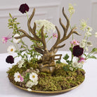 A decorative tabletop arrangement featuring a bronze deer head sculpture with antlers, set in a round gold tray filled with green moss and surrounded by small white, pink, and burgundy flowers on a light surface.