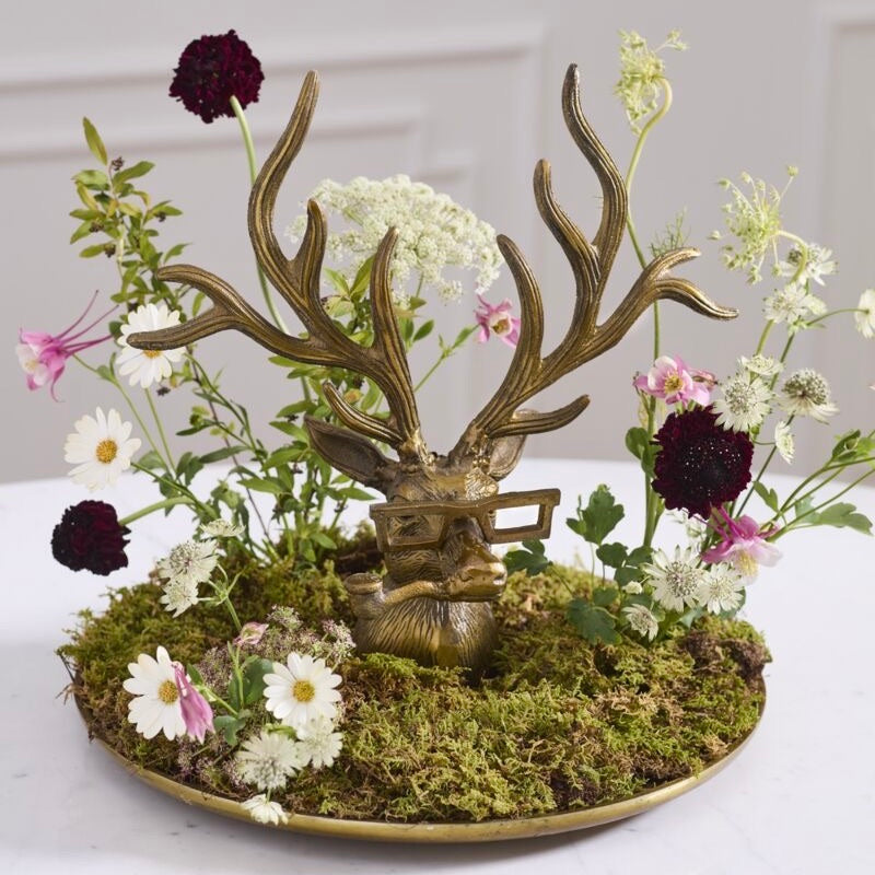A decorative tabletop arrangement featuring a bronze deer head sculpture with antlers, set in a round gold tray filled with green moss and surrounded by small white, pink, and burgundy flowers on a light surface.