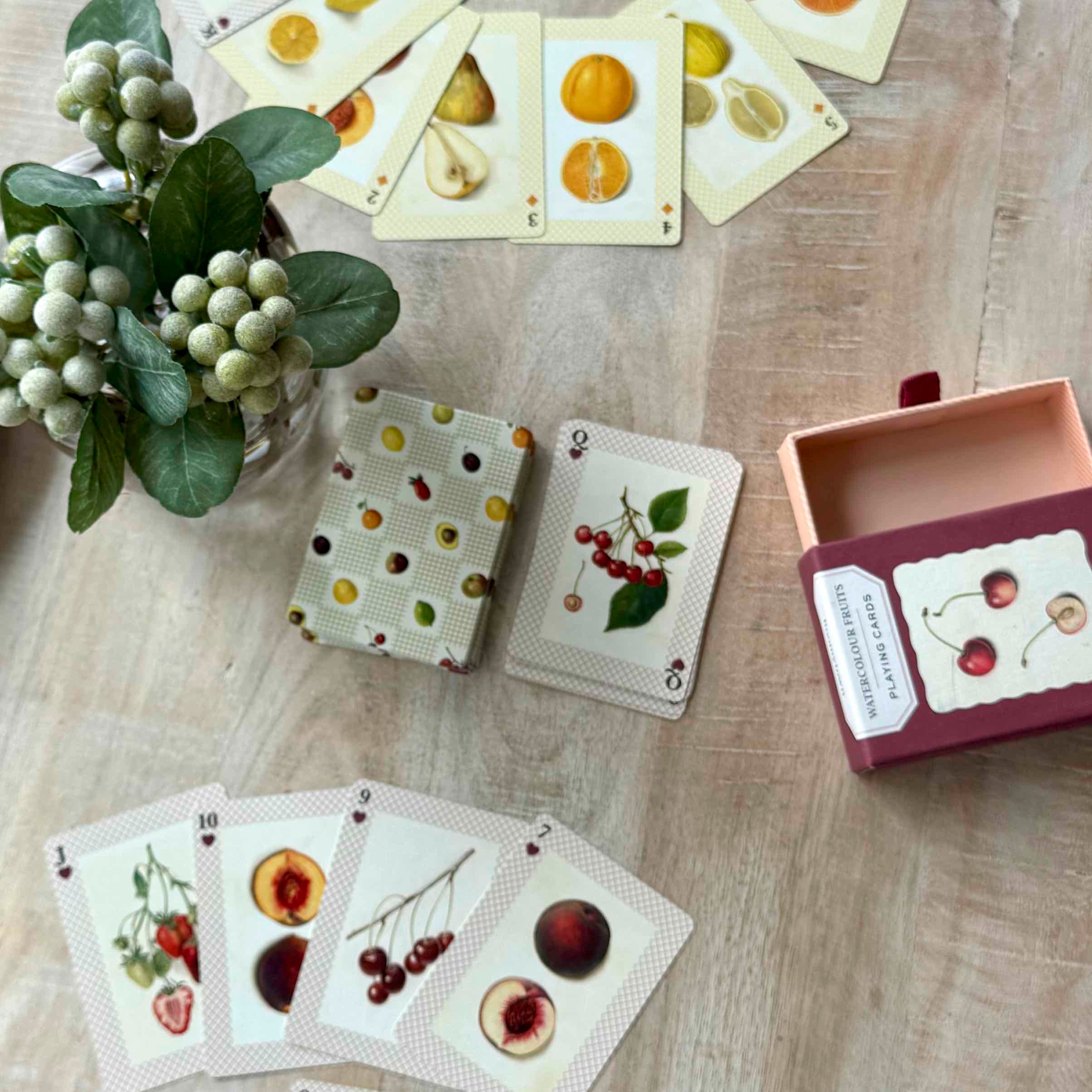 Playing cards with fruit illustrations on a wooden surface, accompanied by a box and decorative plant.