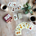 Playing cards with fruit illustrations on a wooden table with coffee cups and a plant.