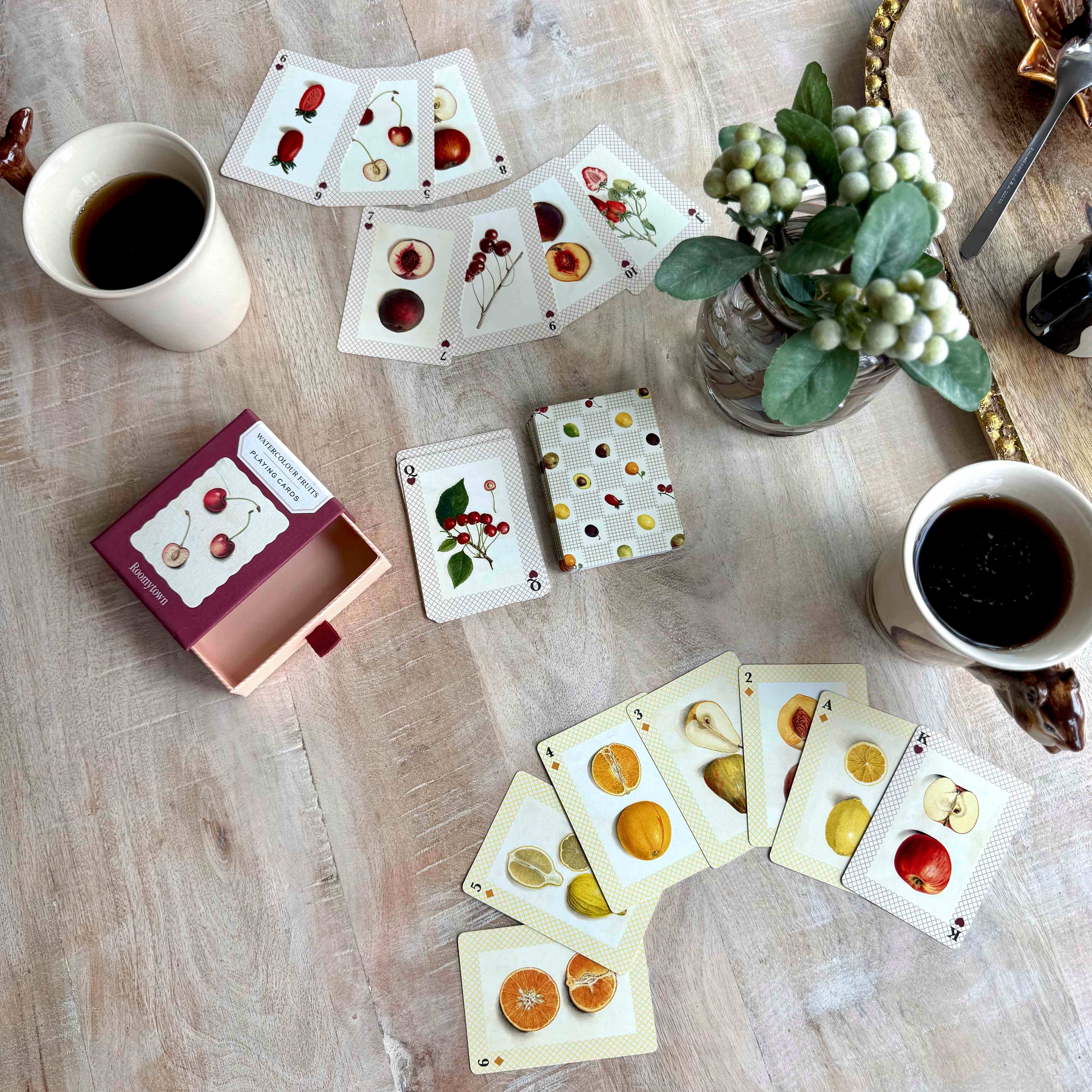 Playing cards with fruit illustrations on a wooden table with coffee cups and a plant.