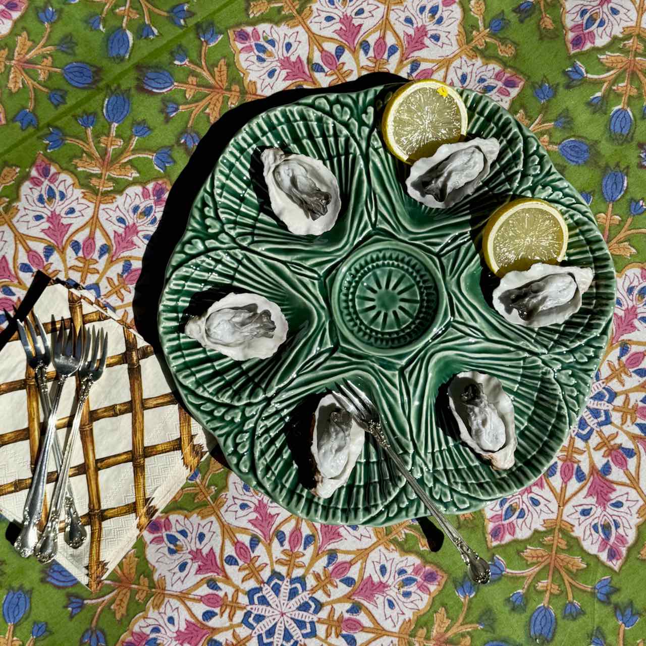 A green ceramic oyster shell platter with six indentations, styled with six raw oysters, two lemon wedges, forks, and a napkin on a vibrant floral tablecloth.